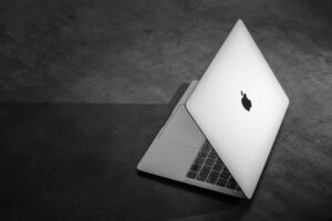 A PARTIALLY-OPENED WHITE APPLE LAPTOP SITS ON A DARK FLOOR.