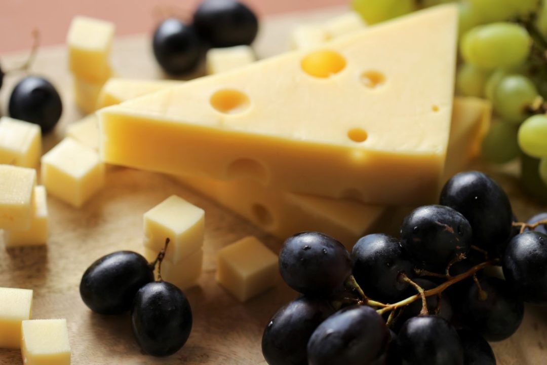 Piece of cheese sits with green and black grapes on a wooden board on brown background.