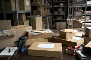 SEVERAL PACKAGES AND A SCANNER SIT ON A CLUTTERED TABLE WITH SHELVES FULL OF PACKAGES IN THE BACKGROUND.