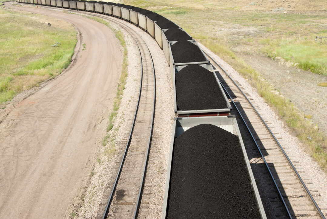 SEVERAL RAIL CARS LOADED WITH COAL ARE BEING TRANSPORTED ON TRACKS SURROUNDED BY DIRT AND SOME GRASS.