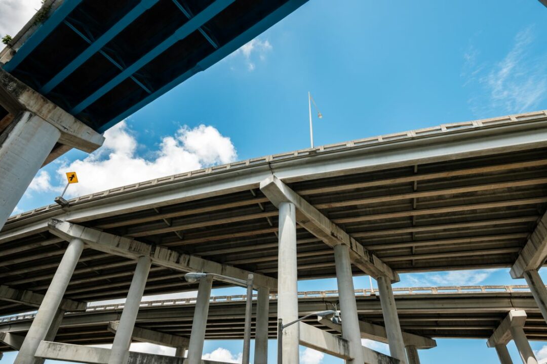 A HIGHWAY OVERPASS, SEEN FROM BELOW, ARCS ACROSS A BLUE SKY