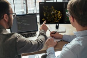 TWO WORKERS DISCUSS DATA SHOWN ON COMPUTER SCREENS