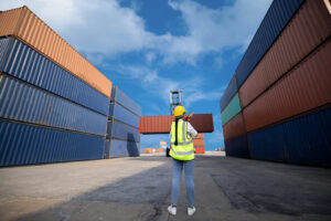 A WOMAN'S BACK IS FACING US. SHE IS IN A CONSTRUCTION WORKER UNIFORM AND MANAGING SHIPPING CONTAINERS.