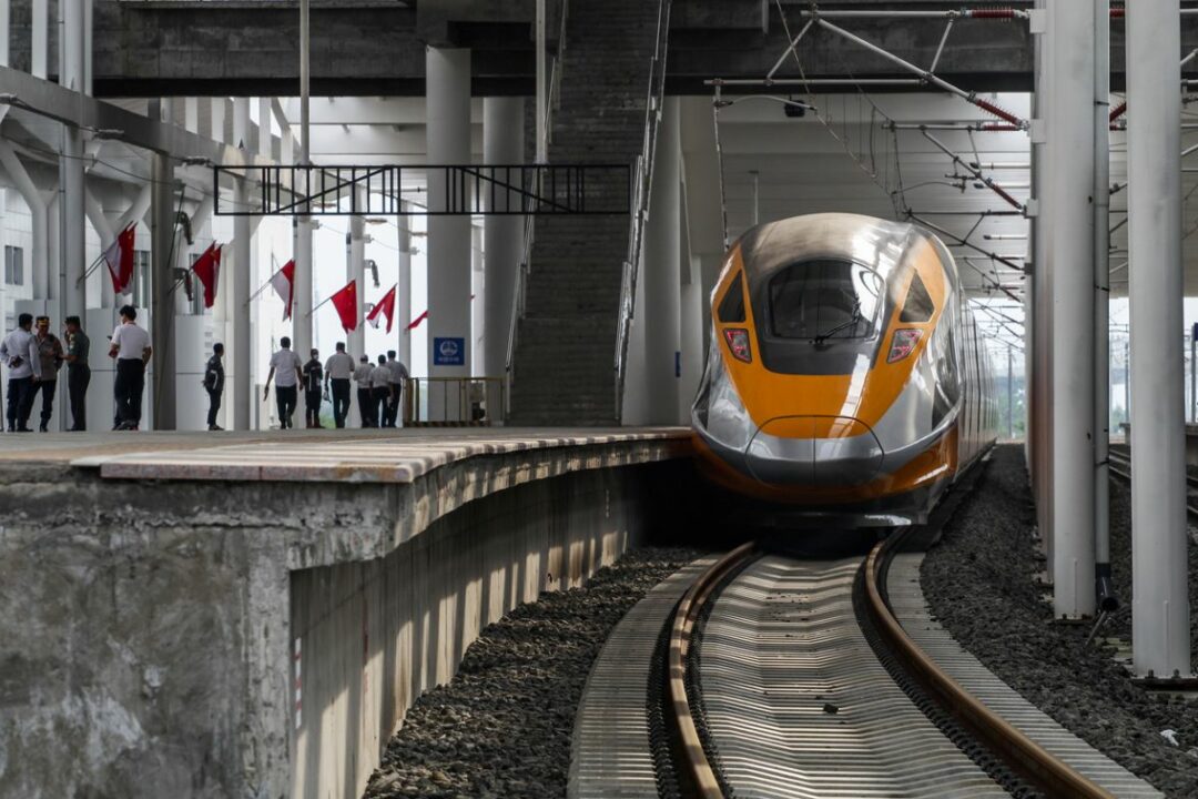 A SLEEK MODERN TRAIN ENTERS ALONGSIDE A CURVING PLATFORM IN A STATION BEDECKED WITH FLAGS