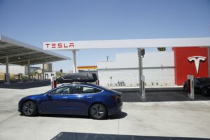 A TESLA MODEL 3 EV CAN BE SEEN LEAVING A TESLA-LABLED CHARGING STATION.