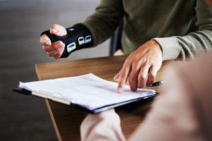 A PERSON WITH A BLACK MEDICAL WRAP ON THEIR HAND IS SITTING AT A DESK WITH ANOTHER PERSON. THE INJURED INDIVIDUAL IS USING THEIR NON-INJURED HAND TO POINT AT A DOCUMENT ON A CLIPBOARD.