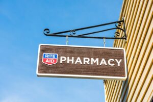 A RITE AID SIGN HANGS OFF THE SIDE OF A BUILDING AGAINST A BRIGHT BLUE SKY