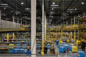 EMPLOYEES IN ORANGE VESTS WORK INSIDE AN AMAZON FULFILLMENT CENTER.