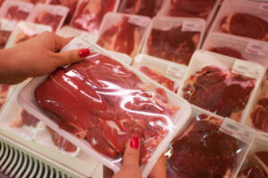 A WOMAN'S HANDS HOLDS PACKAGED MEAT IN FRONT OF A STOCKED SHELF OF PACKAGED MEAT IN A GROCERY STORE.