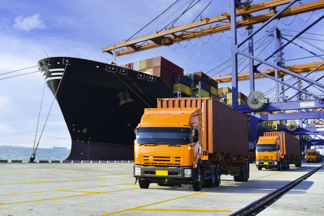 A FLEET OF THREE ORANGE TRUCKS DRIVE ON THE DOCK OF A PORT NEXT TO A LARGE CONTAINER SHIP.