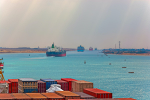 LARGE CONTAINER SHIPS TRAVEL ON THE SUEZ CANAL WITH CONTAINERS IN THE FOREGROUND OF THE PHOTO.