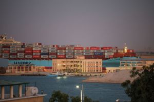 A HUGE CONTAINER SHIP PUSHES THROUGH A NARROW WATERWAY, BUILDINGS VISIBLE IN THE FOREGROUND