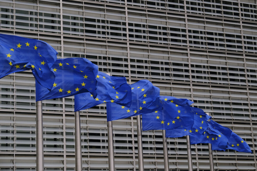 SEVERAL EU FLAGS ON FLAGPOLES FLAPPING IN THE WIND IN FRONT OF A LARGE OFFICE BUILDING.