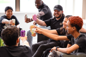 A GROUP OF SMILING SEATED PEOPLE RAISE SOFT DRINKS CANS TO EACH OTHER