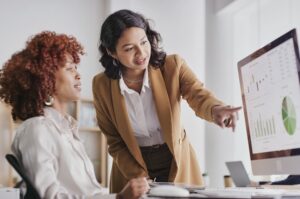 A WOMAN STANDS WHILE ANOTHER WOMAN SITS, BOTH LOOKING AT A COMPUTER SCREEN WITH CHARTS ON IT.