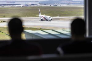 TWO PEOPLE IN SILHOUETTE WATCH A PLANE ON AN AIRPORT TARMAC FROM THE TERMINAL WINDOW.jpg