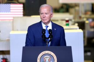 AN ELDERLY MAN IN A BLUE SUIT STANDS BEHIND A PODIUM LABELED PRESIDENT OF THE UNITED STATES