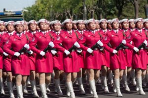HUNDREDS OF WOMEN IN RED MILITARY UNIFORMS MARCH IN LOCKSTEP DOWN A TREE-LINED AVENUE