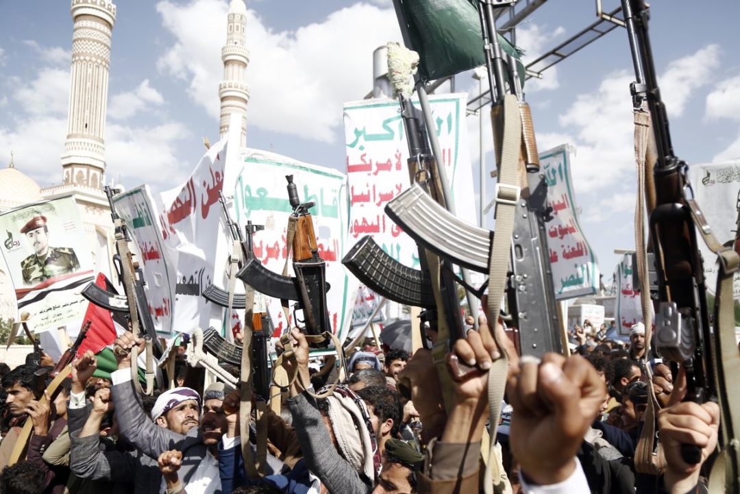 A TEEMING CROWD OF MEN HOLD ALOFT AUTOMATIC WEAPONS AGAINST A BACKDROP OF ARABIC BANNERS AND MINARETS.