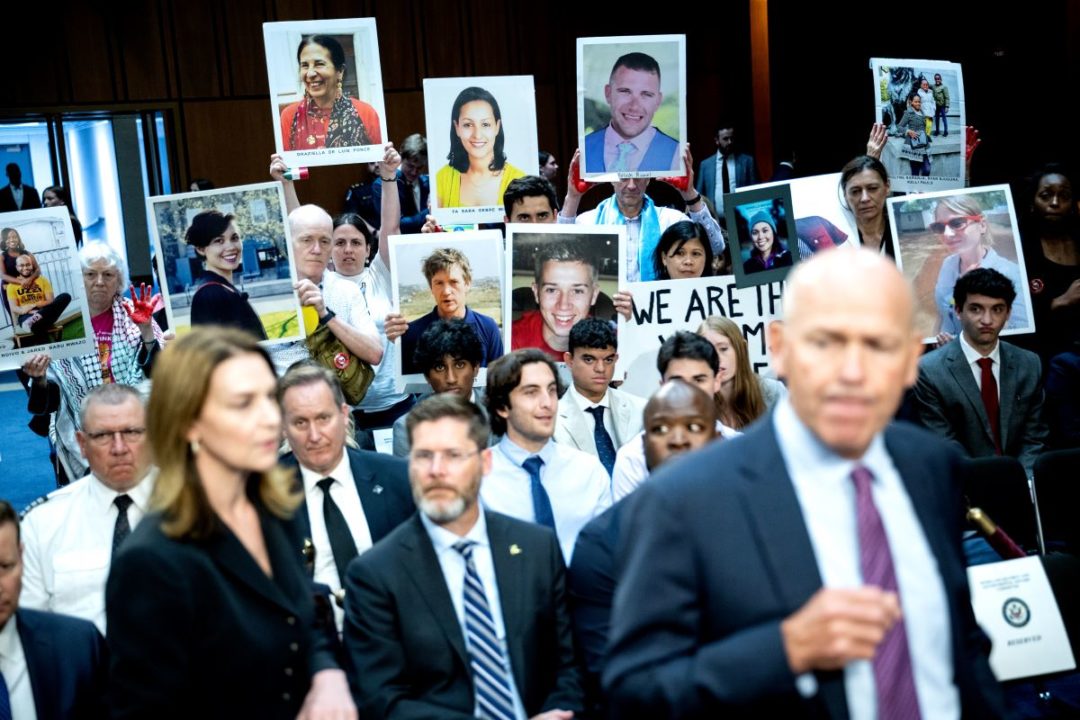 A BALDING MIDDLE AGED MAN IN A SUIT HURRIES PAST PEOPLE HOLDING PHOTOS OF RELATIVES