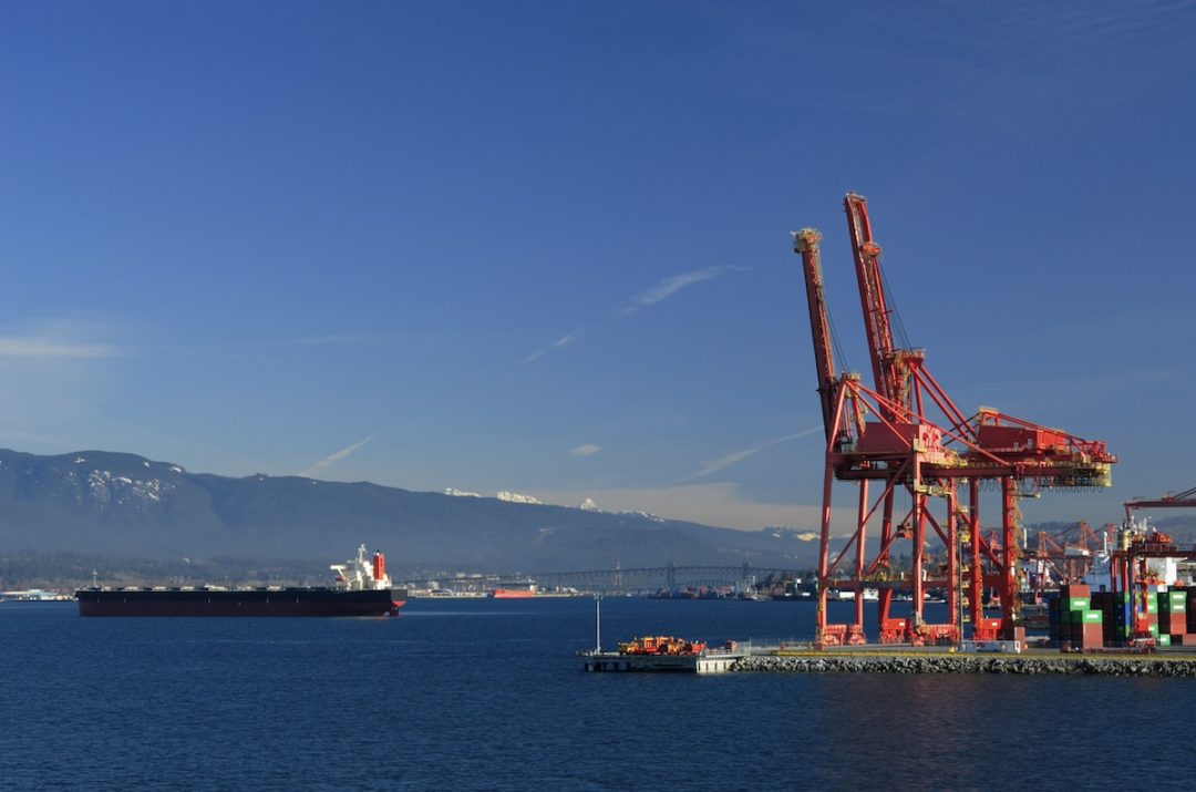 Two large red cranes at a shipping port, with a black container ship moving past the shore.