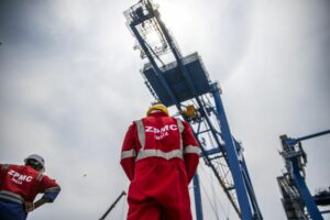 MEN WEARING BRIGHT RED SAFETY SUITS EMBLAZONED WITH ZPMC INDIA STAND IN FRONT OF A GIANT CRANE FRAMED BY A CLOUDY SKY