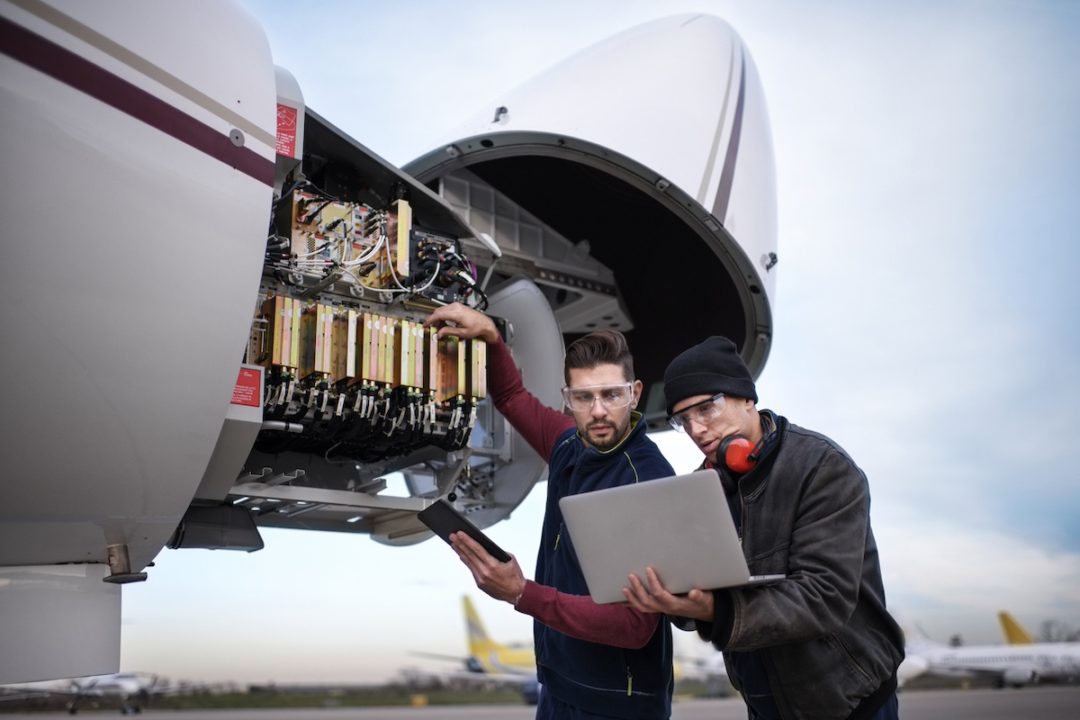 A mechanic next to the opened nose of an airplane, replacing electrical components while holding a laptop