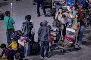 A LONG RAMBLING LINE OF PEOPLE STAND WITH LUGGAGE CARTS IN AN AIRPORT TERMINALERG.jpg