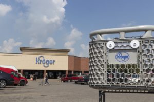 A grocery cart with the Kroger logo on it, with the front of a Kroger grocery store in the background