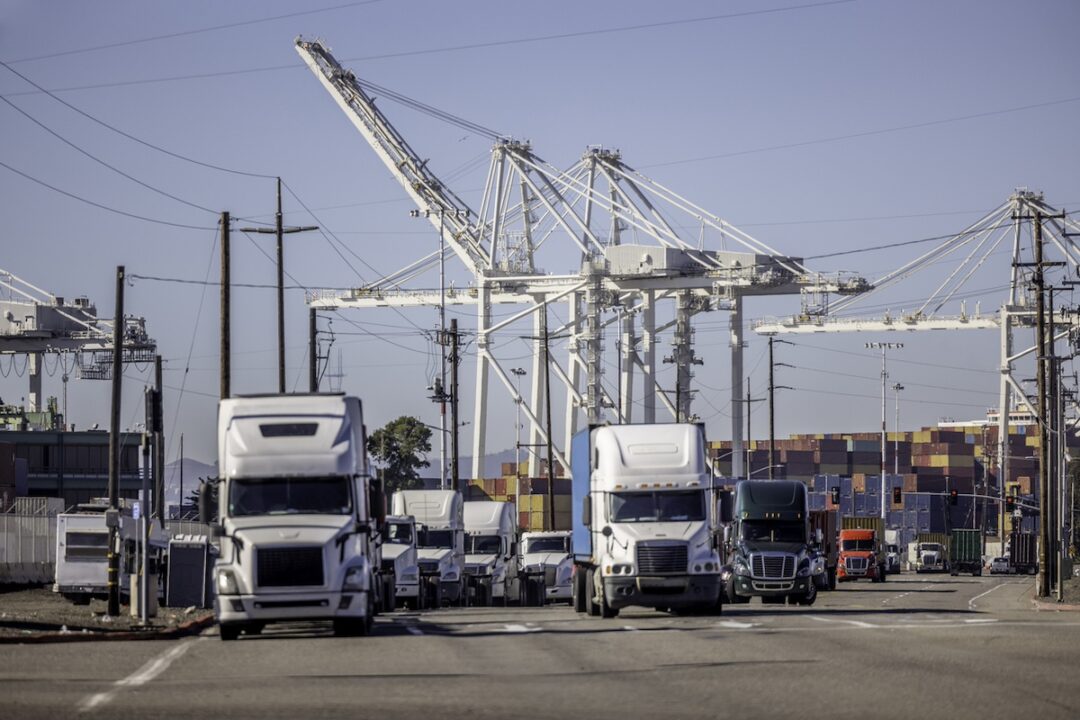 A front-facing view of a line of semi-trucks towing shipping containers, with two white shipping cranes in the background