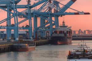 A line of blue shipping cranes unloading a container ship at sunset