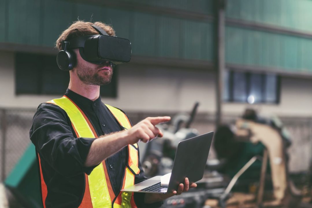 A WORKER WEARING A HI-VIS VEST AND VIRTUAL REALITY GOGGLES POINTS AT A SPOT JUST ABOVE AN OPEN LAPTOP
