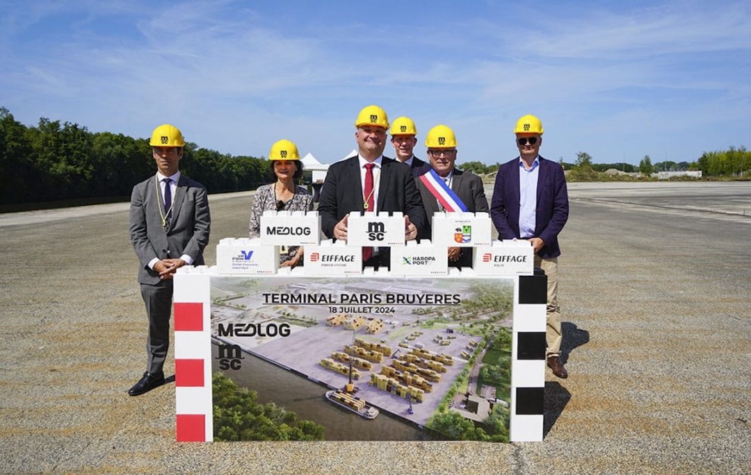 Five men in suits and one woman in a grey dress, all wearing yellow construction hats and standing in front of an artist rendering of a new shipping terminal