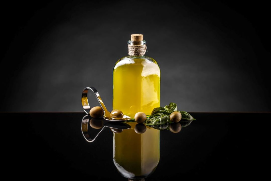 A BOTTLE OF OLIVE OIL STANDS SURROUNDED BY OLIVES ON A BEAUTIFUL DARK REFLECTIVE SURFACE