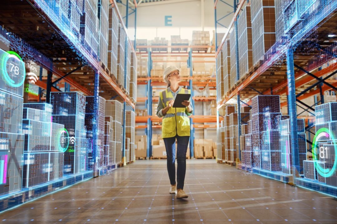 A WORKER IN HI-VIS VEST AND HARDHAT WALKS DOWN A WAREHOUSE AISLE CONSULTING A TABLET COMPUTER