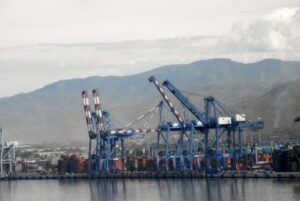 A row of blue shipping cranes, above stacks of orange shipping containers along a coastline