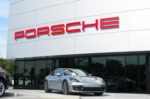 A grey sports car parked outside of a Porsche dealership, with the carmaker's brand written in red on the outside of the building