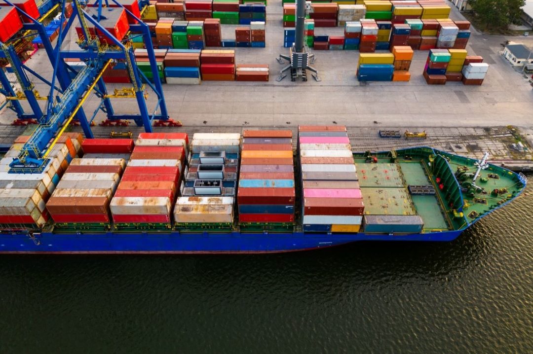 A LARGE CONTAINER SHIP, SEEN FROM ABOVE, IS LOADED WITH MULTI-COLORED CONTAINERS AT DOCK