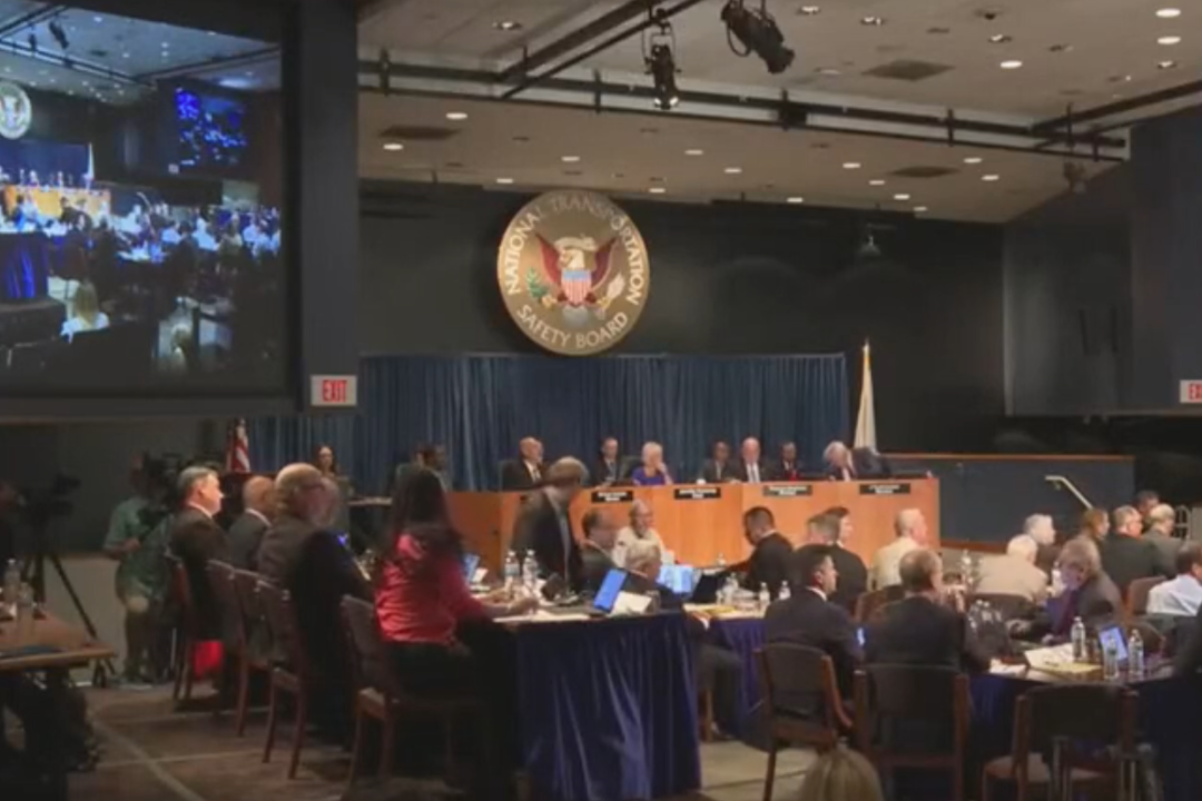 A wide shot of a room full of people sitting in front of a dais under an NTSB logo, aside a large projection screen