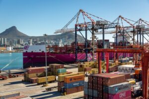 Rows of multi-colored containers stacked up in a shipyard, with a red container vessel docked beneath three large cranes