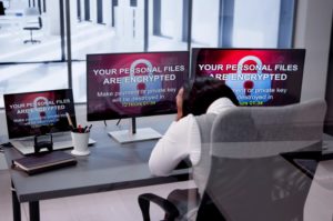 AN OFFICE WORKER HOLDS HER HEAD IN HER HANDS IN FRONT OF 3 SCREENS THAT ALL BEAR THE MESSAGE: YOUR PERSONA FILES ARE ENCRYPTED