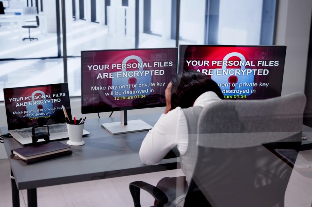 AN OFFICE WORKER HOLDS HER HEAD IN HER HANDS IN FRONT OF 3 SCREENS THAT ALL BEAR THE MESSAGE: YOUR PERSONA FILES ARE ENCRYPTED