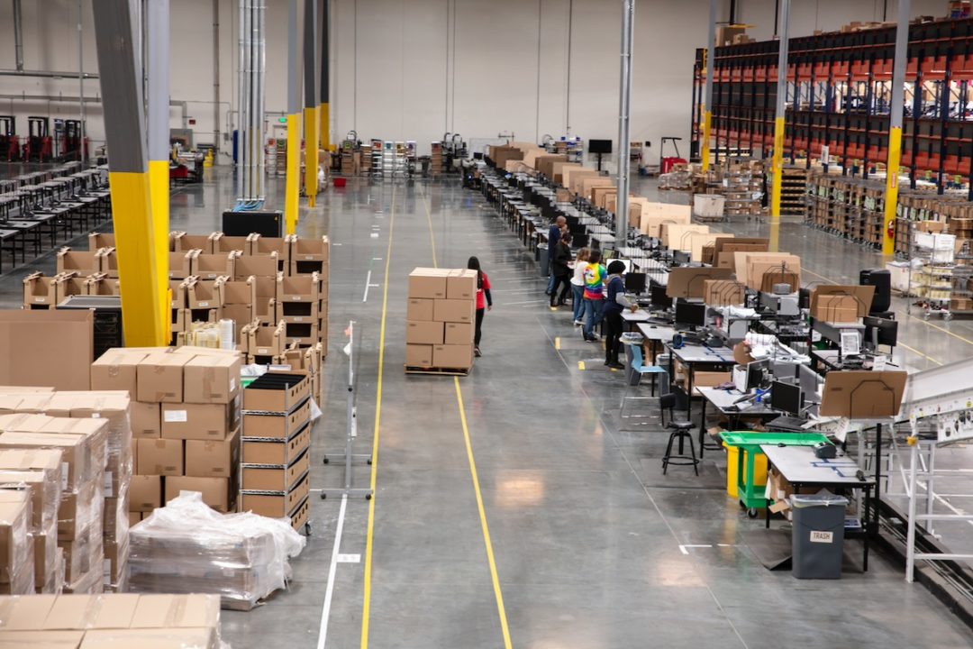 A wide shot of a row of cardboard boxes arranged along a warehouse floor, with works standing at desks alongside the boxes