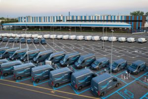 A parking lot full of navy blue and white Amazon delivery vans, with a striped blue and white building in the background.