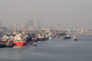 A rec container ship docked at a shipping port, set against a hazy downtown city skyline