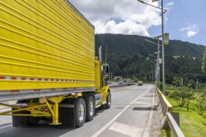 A yellow semi-truck driving on a road, set against a green hillside