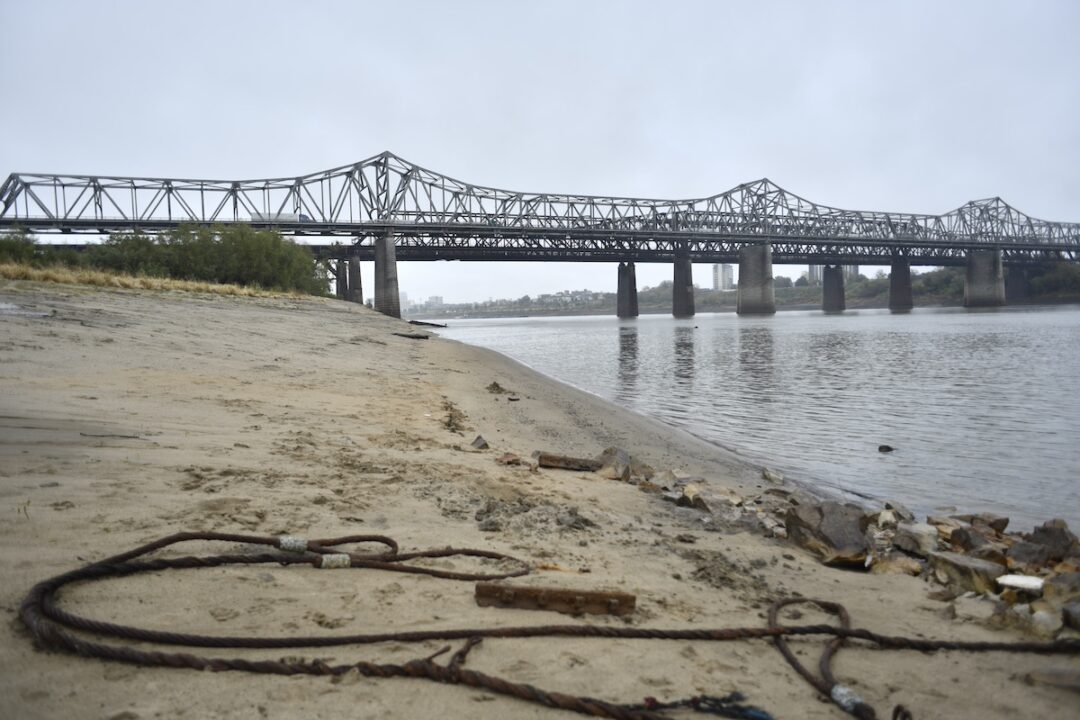 A sandy shoreline along a river with a bridge in the background