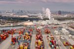 Rows of stacked, multi-colored shipping containers in a shipyard, with a collection of white industrial buildings in the background