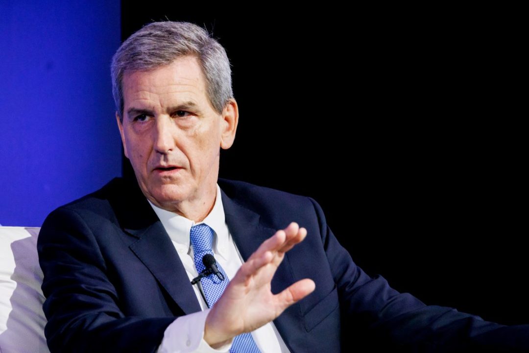 A GREY-HAIRED MAN IN A NAVY BLUE SUIT AND LIGHT BLUE TIE RAISES HIS RIGHT HAND IN AN EMPHATIC GESTURE