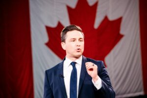 A MAN IN A DARK BLUE SUIT AND TIE IN FRONT OF A CANADIAN FLAG PINCHES HIS FINGER AND THUMB TOGETHER IN A GESTURE